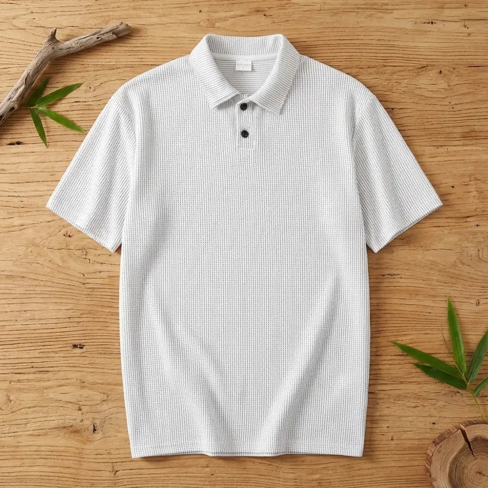 Polo homme en maille de bambou blanc posé à plat sur une table en bois brut avec feuilles de bambou