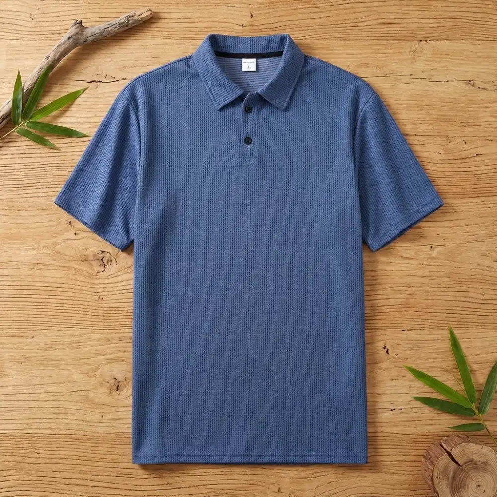 Polo homme en maille de bambou bleu posé à plat sur une table en bois brut avec feuilles de bambou