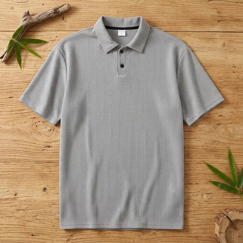 Polo homme en maille de bambou gris posé à plat sur une table en bois brut avec feuilles de bambou