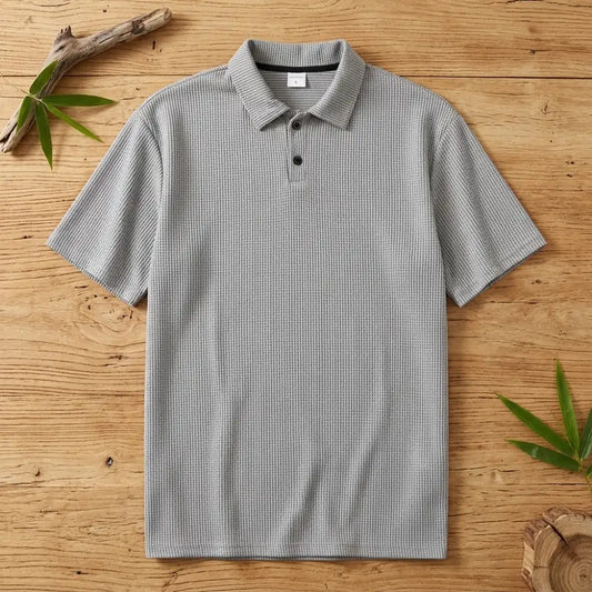 Polo homme en maille de bambou gris posé à plat sur une table en bois brut avec feuilles de bambou