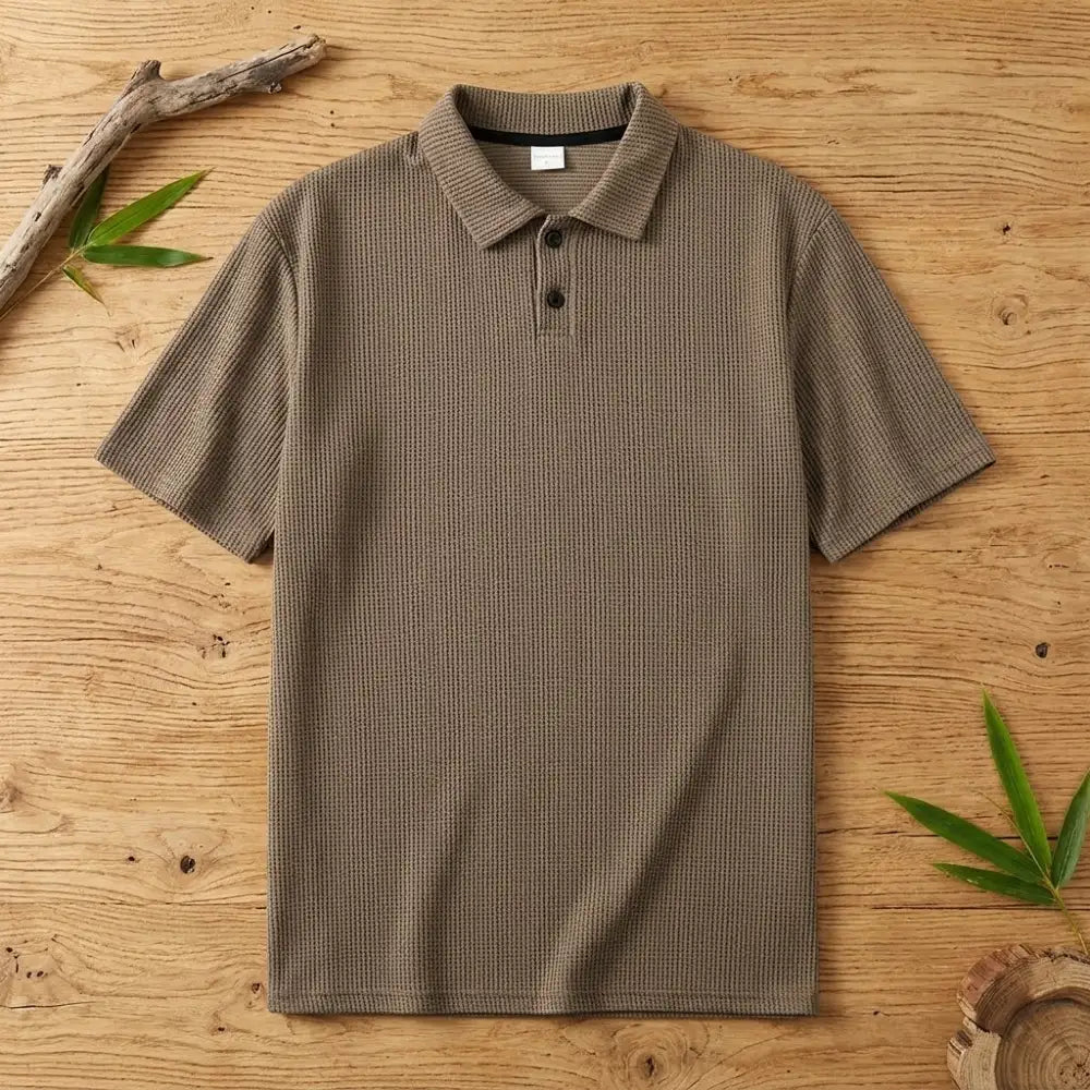 Polo homme en maille de bambou marron posé à plat sur une table en bois brut avec feuilles de bambou