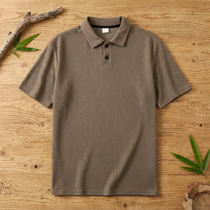 Polo homme en maille de bambou marron posé à plat sur une table en bois brut avec feuilles de bambou