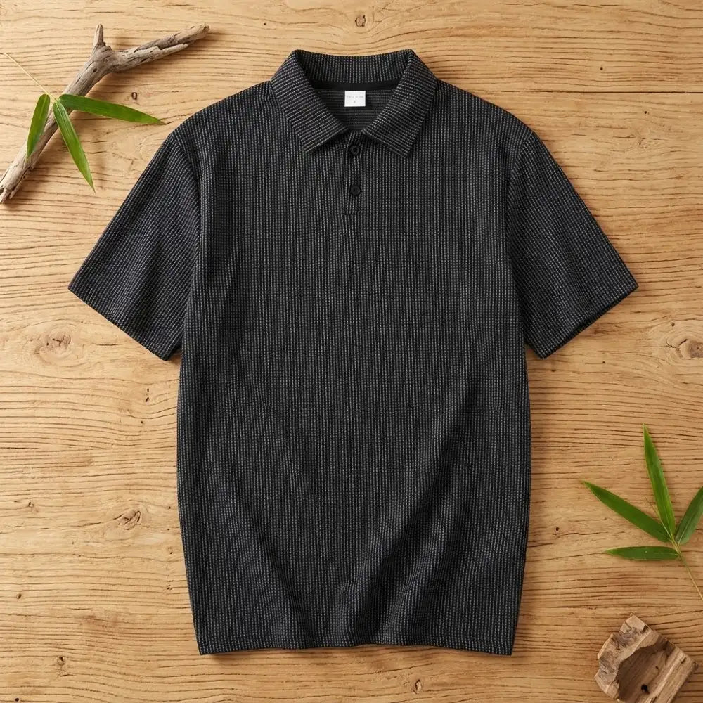 Polo homme en maille de bambou noir posé à plat sur une table en bois brut avec feuilles de bambou