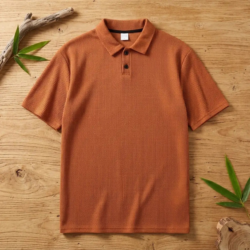 Polo homme en maille de bambou orange posé à plat sur une table en bois brut avec feuilles de bambou