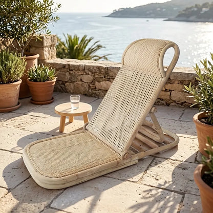 Transat bas en rotin naturel, dossier en cannage et assise tressée, sur une terrasse en pierre face à la mer